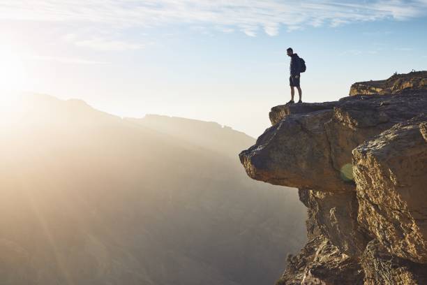 KIITEs On the Edge 1 Young tourist with backpack standing on the edge of cliff at sunrise. Jebel Akhdar, Grand Canyon of Oman.